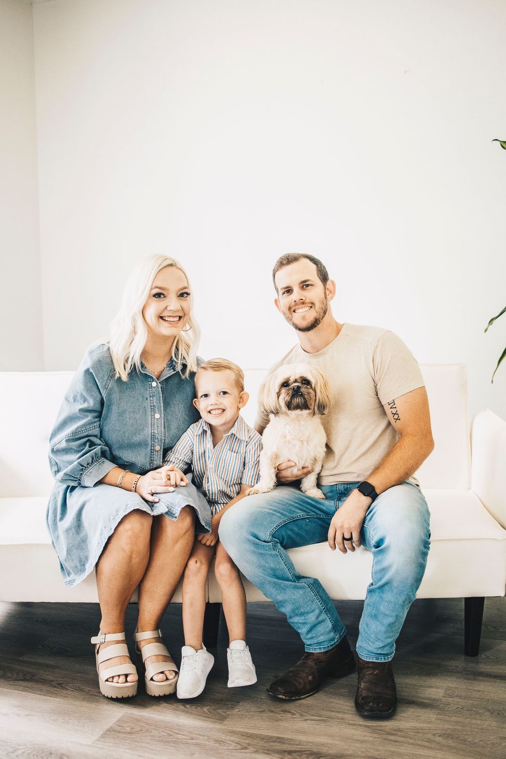 Happy family with blonde mom, young son, bearded dad, and small dog posing on white couch.