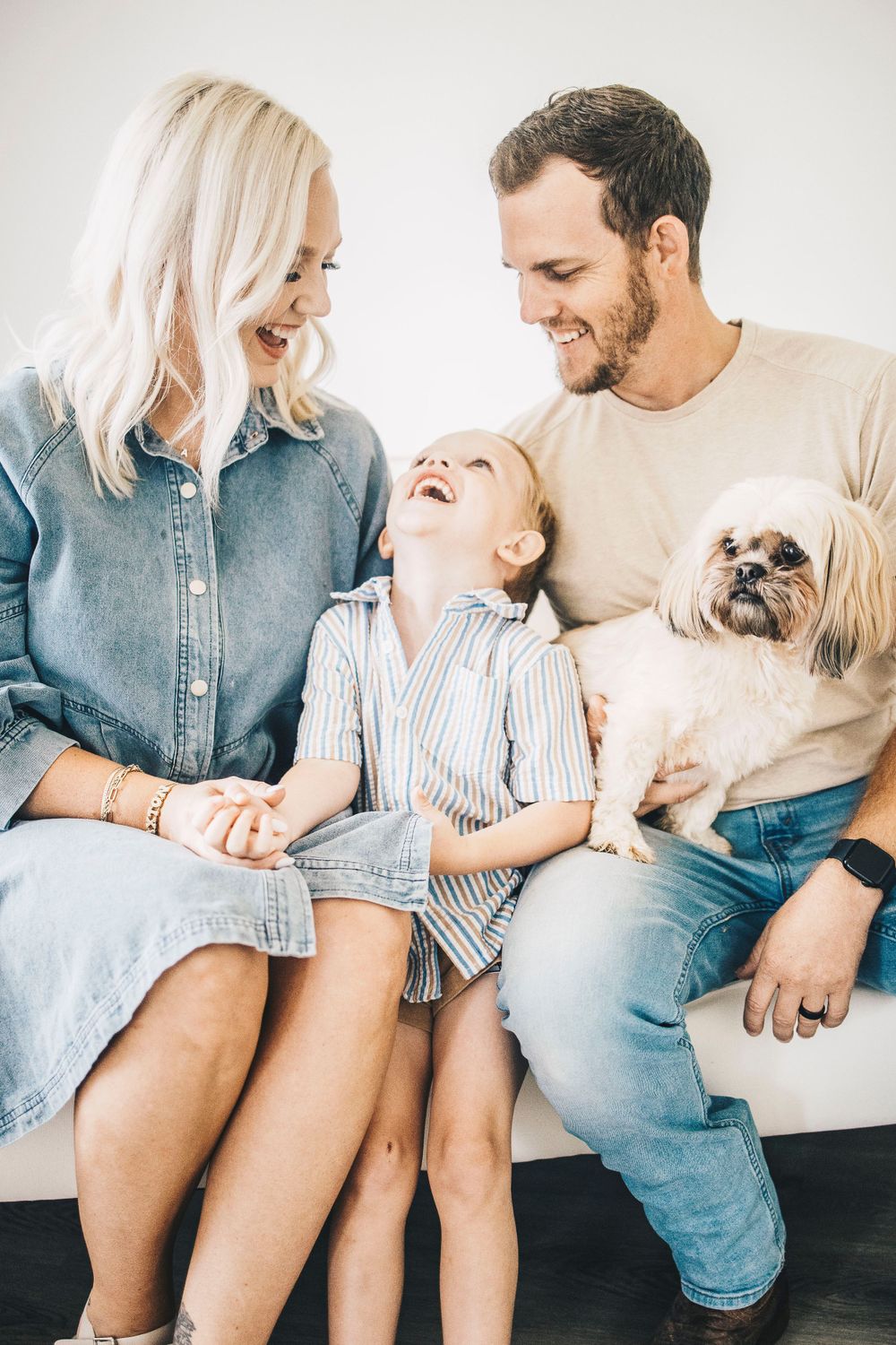Joyful family moment with parents laughing as young boy tilts head back on white couch with small dog.