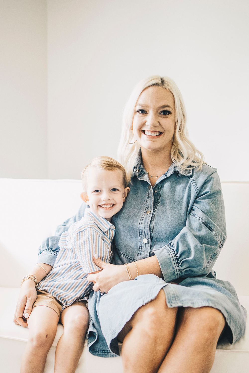 Blonde mother in denim dress smiling with young son in striped shirt on white sofa.
