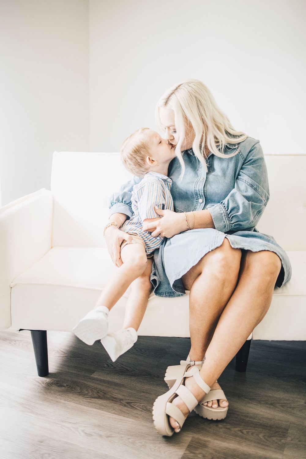 Tender moment of young boy leaning in to kiss blonde mother on white couch indoors.