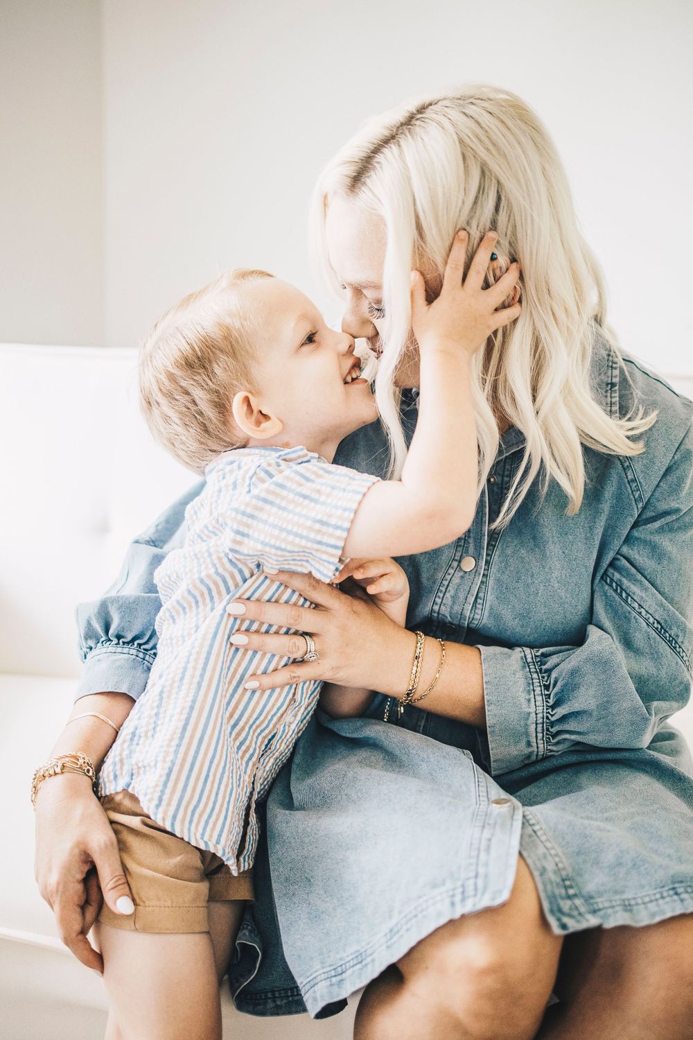 Playful toddler boy touching blonde mothers face while sitting together on white sofa.