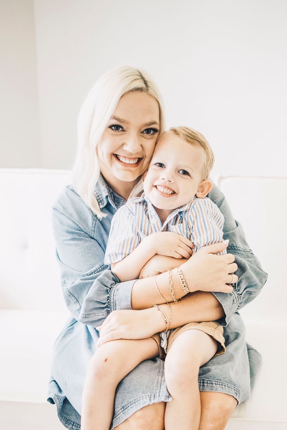Smiling blonde mother hugging laughing toddler boy in striped shirt on white couch.