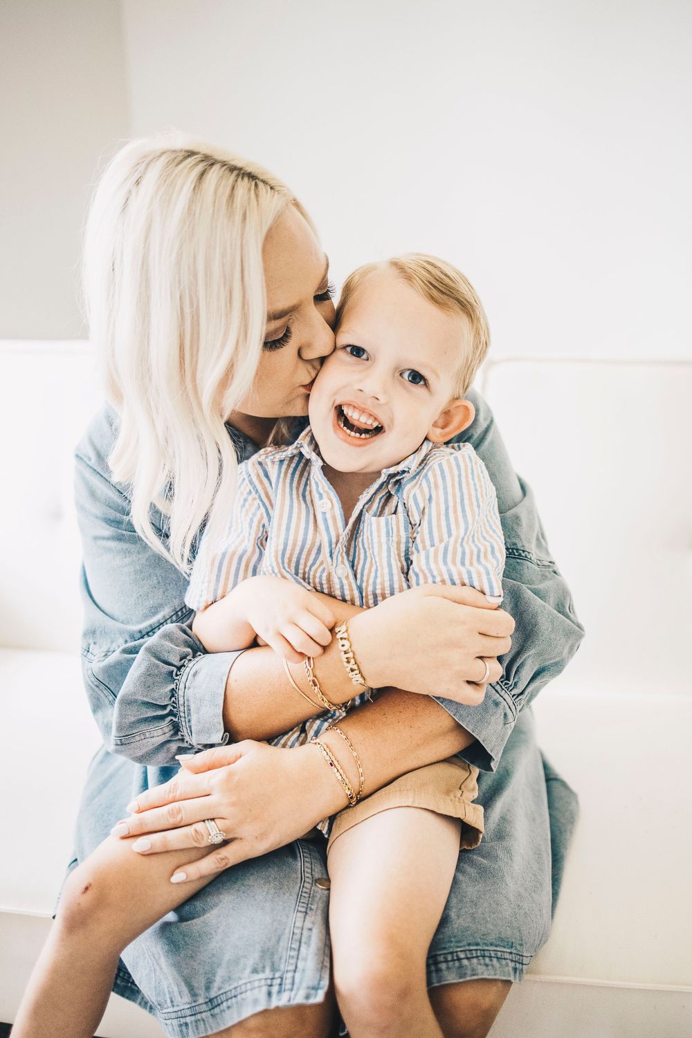 Blonde mother kissing cheek of giggling toddler son while embracing him on white sofa.