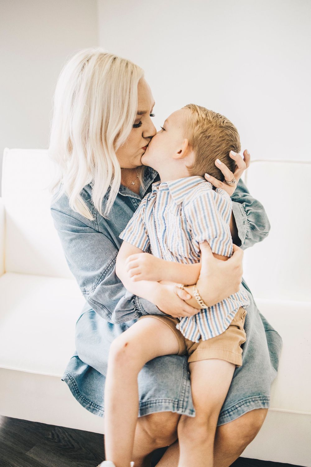 Blonde mother and young toddler son sharing a sweet kiss on white couch indoors.
