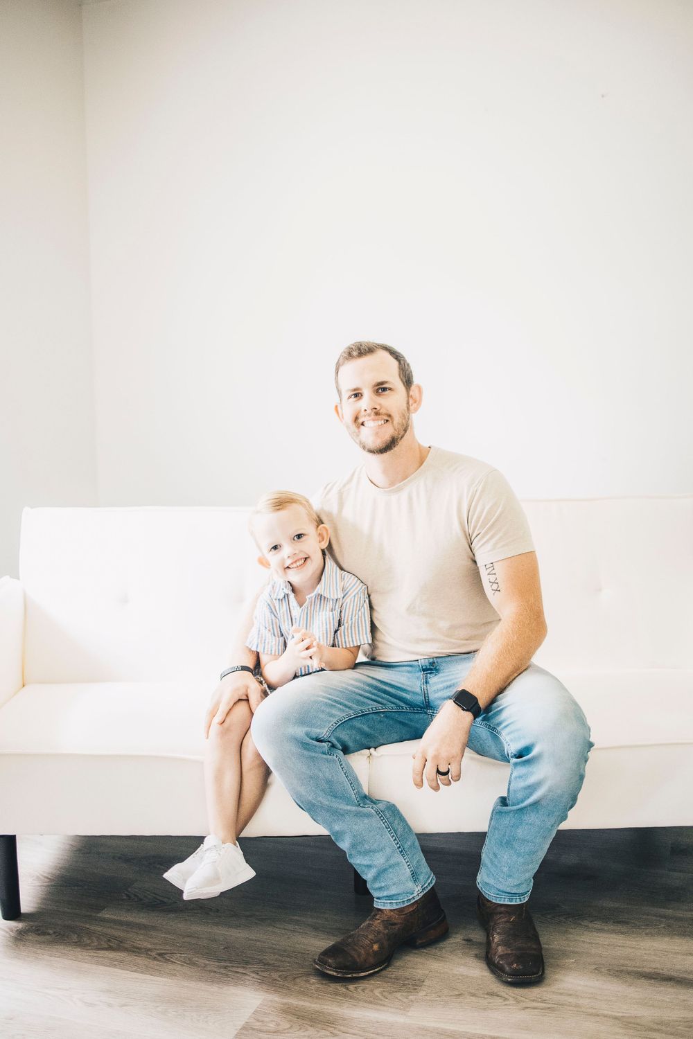 A smiling father and young blonde son sit together on a white couch in a bright room.