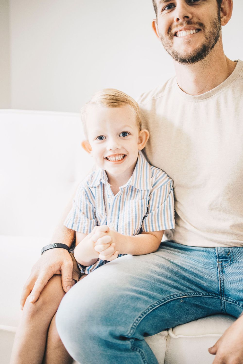 Close-up of a happy blonde toddler boy smiling while sitting with his father on a white sofa.