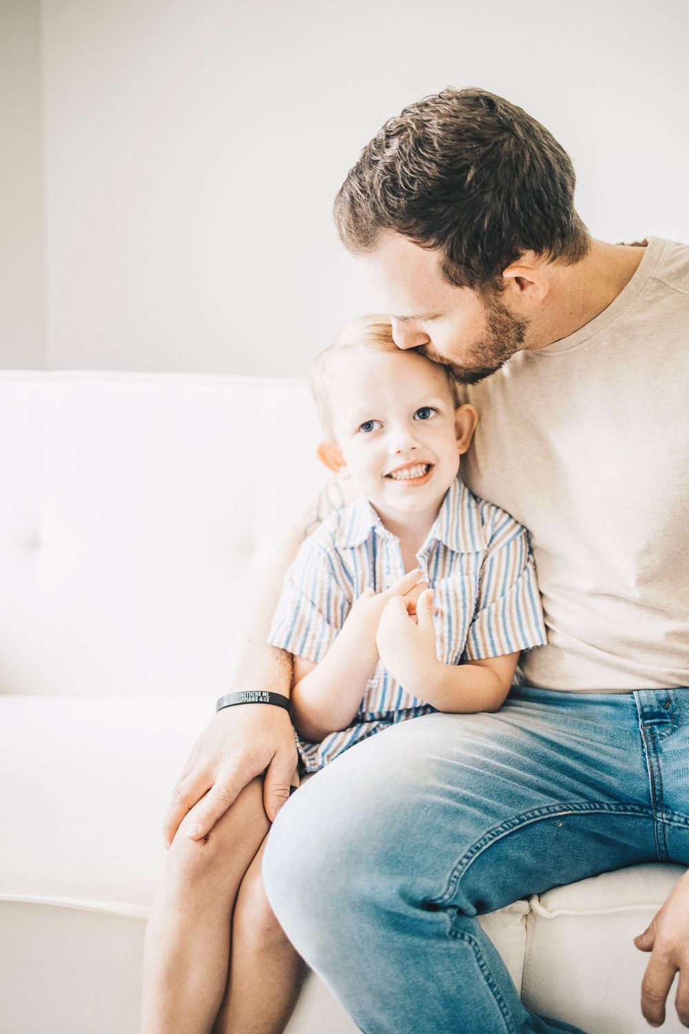 A bearded father kisses his smiling young blonde son on the head while sitting on a white couch.
