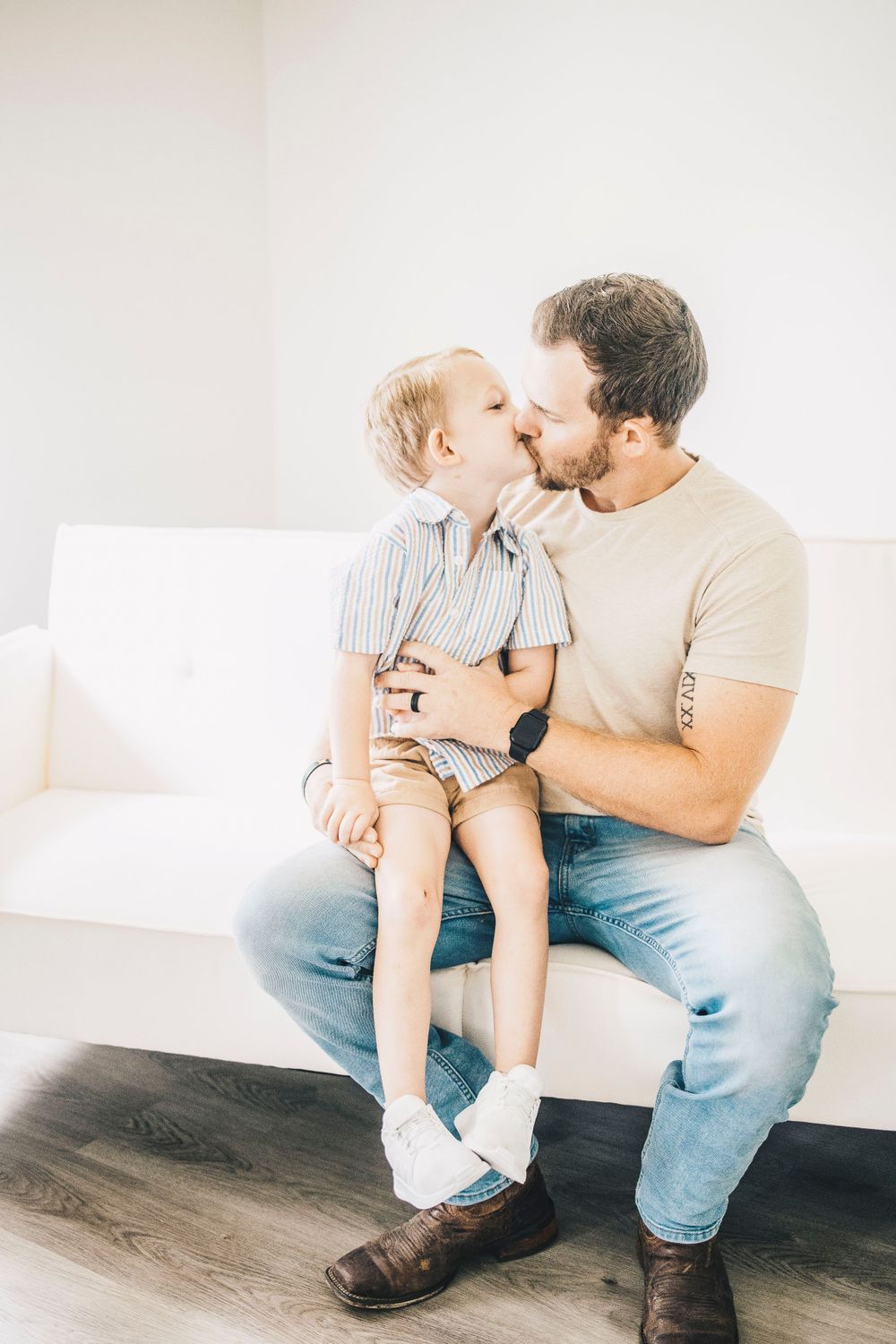 A father and his young blonde son share a sweet kiss while sitting on a white couch.