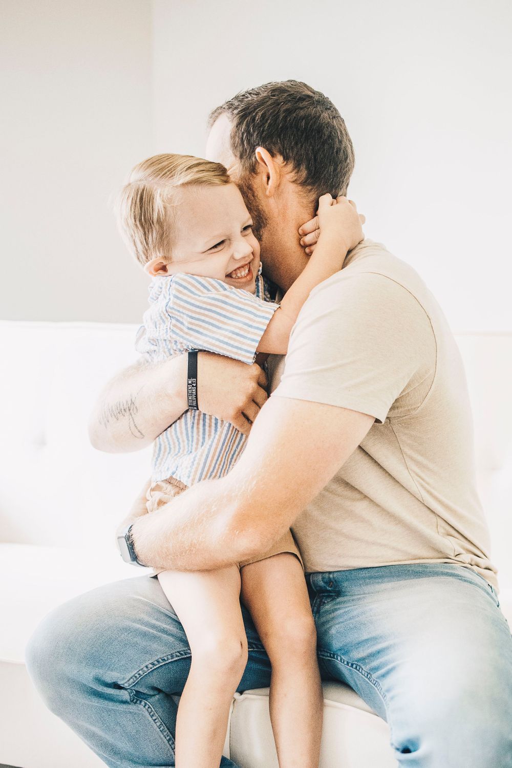 A laughing toddler boy hugs his father tightly while being held on a bright white couch.