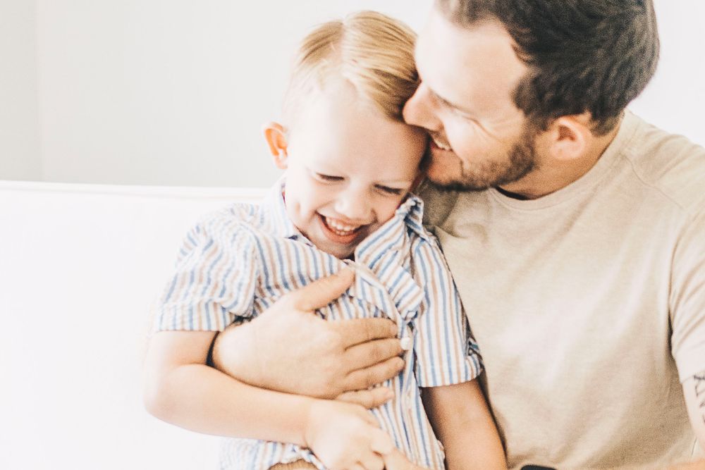 A father cuddles and kisses his giggling young blonde son in a bright white room.