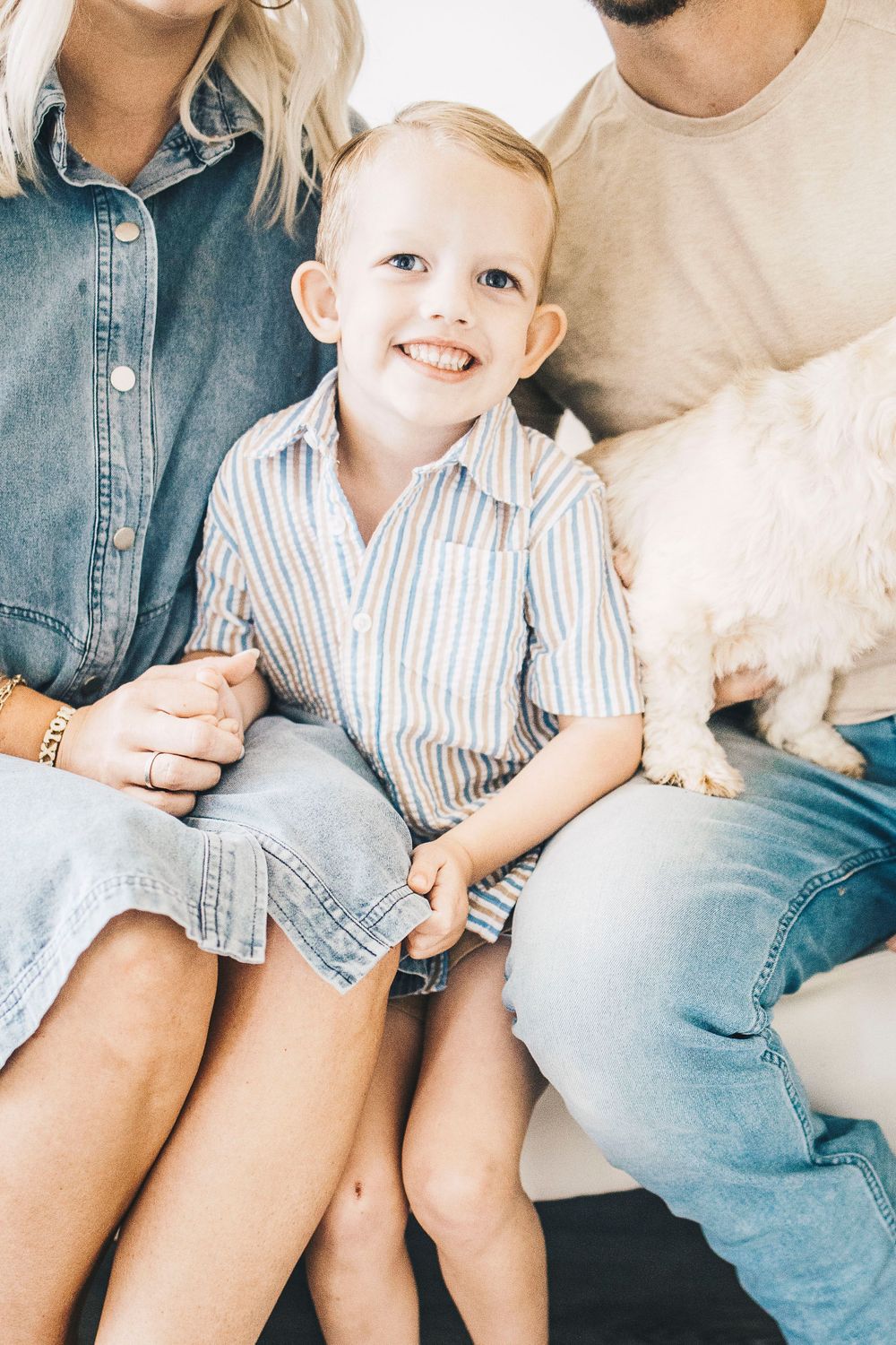 Close-up of smiling toddler boy in striped shirt sitting between parents on white sofa with dog nearby.
