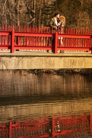 Romantic couple on red bridge over water with beautiful reflection during sunset.