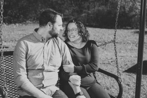 A couple sits together on a swing set in a casual black and white portrait.