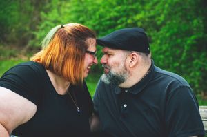 A couple in black clothing sharing an intimate moment outdoors with lush green foliage in the background.