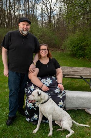 A couple and their white pit bull dog pose together on a wooden bench in a green spring setting.
