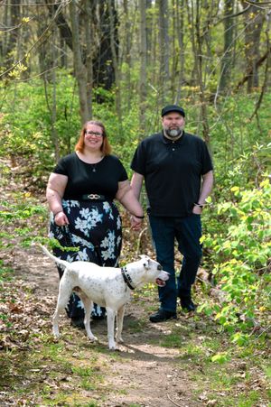 A white dog stands on a forest trail with two people surrounded by lush green foliage on a sunny day.