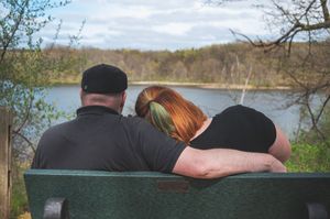 Three people sitting together on a wooden bench overlooking a peaceful lake with autumn trees in the background.