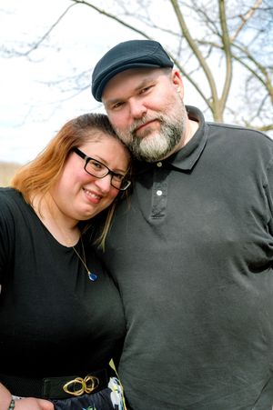 A couple embraces outdoors on a sunny day while wearing casual clothes including a black hat and glasses.
