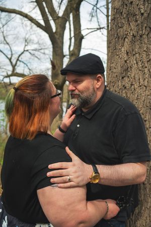 A couple in black clothing shares a tender moment together while standing next to a large tree in a park.