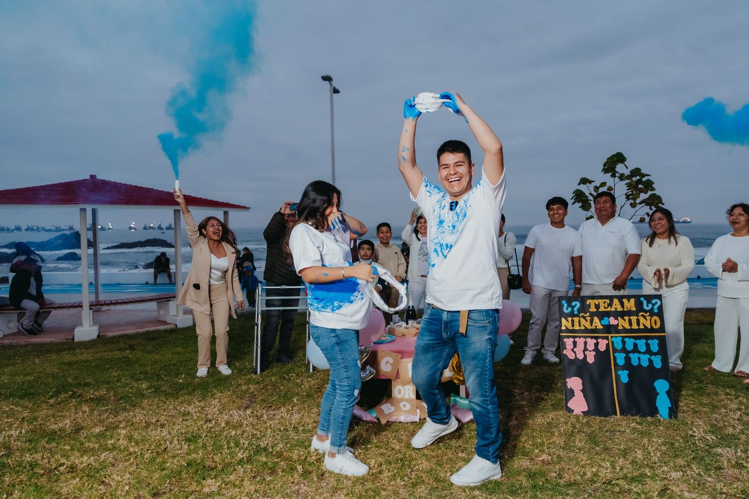Group celebrating with blue smoke bombs and signs outdoors during gender reveal party.