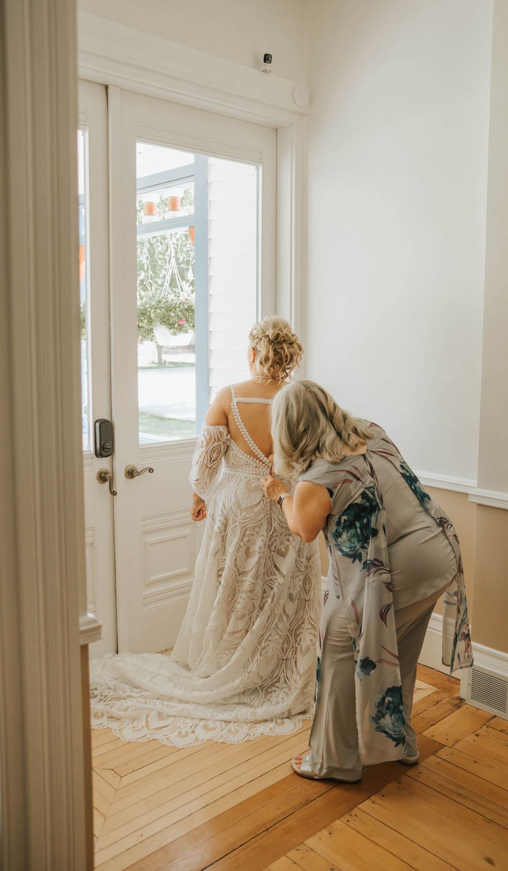 A bride in a flowing lace wedding gown being helped with final preparations in a bright room.