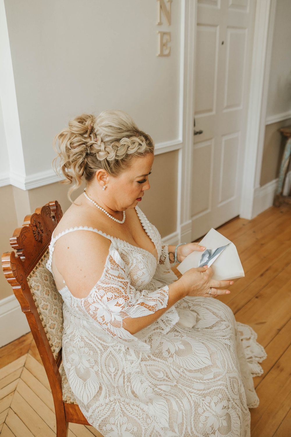 A bride in a pearl-adorned lace gown sits in a wooden chair examining wedding details.