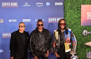 Three attendees pose at Super Bowl event wearing dark clothing against branded backdrop.