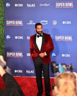 A person in a velvet burgundy tuxedo jacket poses on red carpet at Super Bowl celebration.