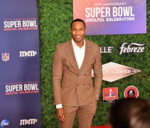Attendee wearing brown checkered suit poses at Super Bowl event with hedge wall backdrop.