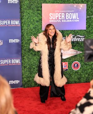 A guest in furry beige coat poses playfully on red carpet at Super Bowl event.