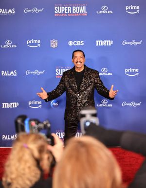 Guest in shimmering black blazer gestures to photographers at Super Bowl event.