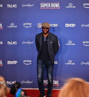A guest in casual attire and newsboy cap poses at Super Bowl event with sponsor backdrop.