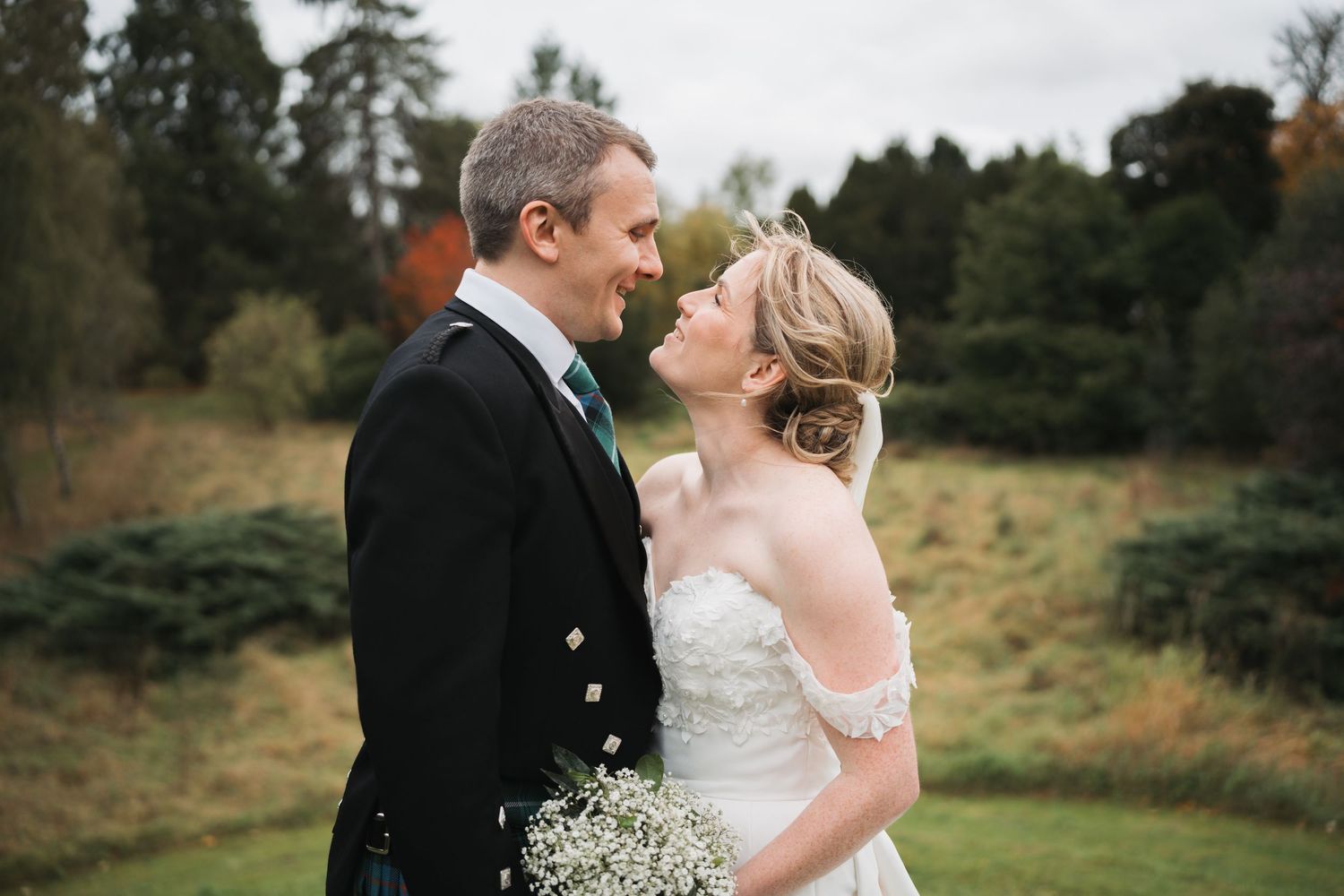 Couple in wedding attire pose on cobblestone street with groom in traditional Scottish kilt.