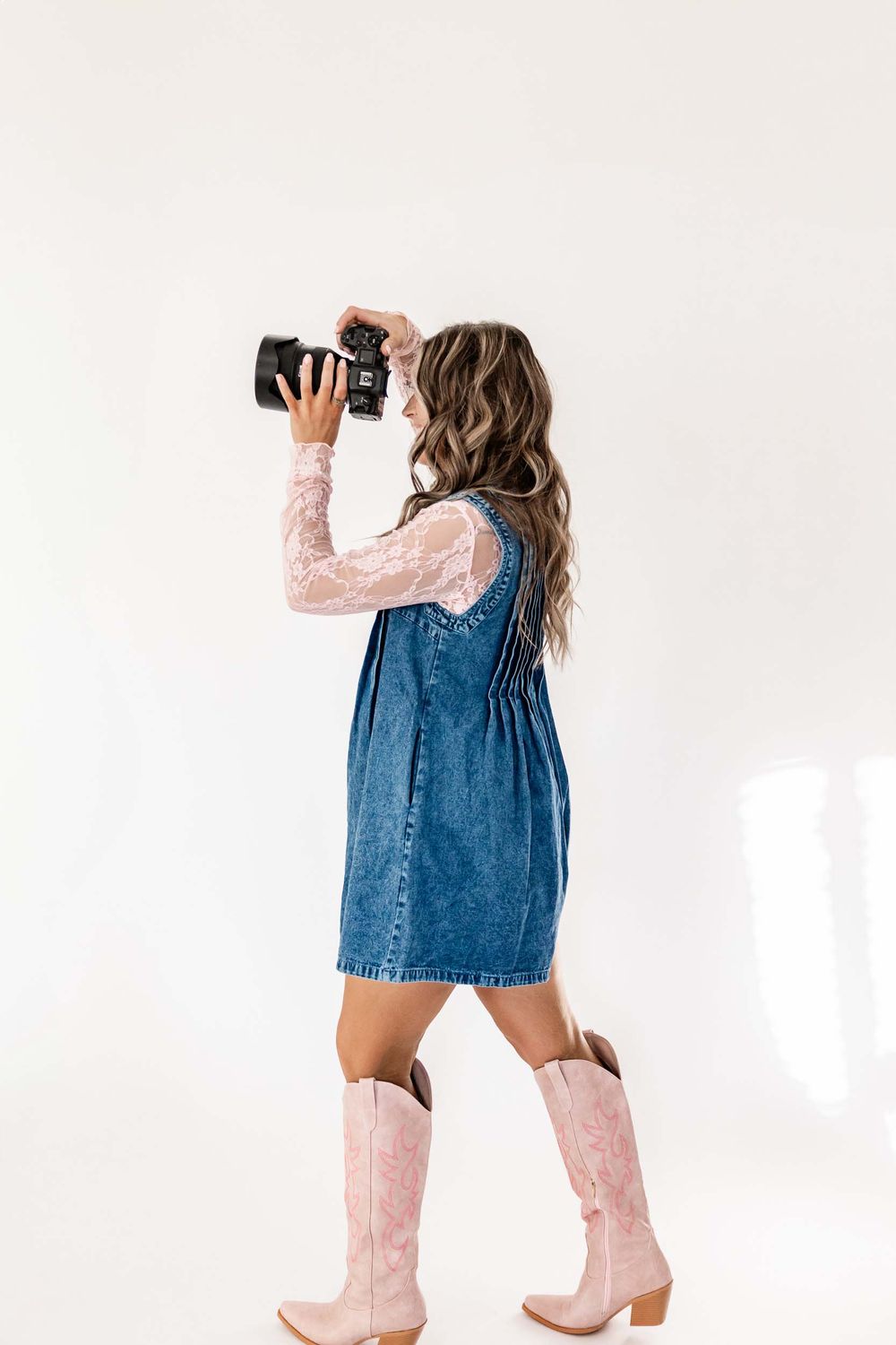 Wyoming Photographer
A photographer in a denim dress and pink cowboy boots holds up a camera while standing against a white backdrop.