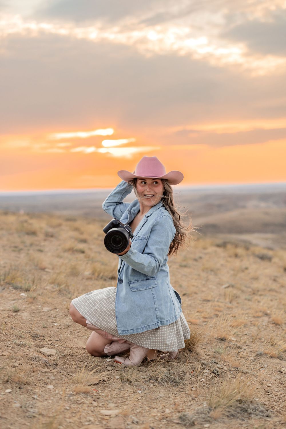 Woman in pink cowboy hat holding camera kneels on dry grass at golden sunset in open landscape.
