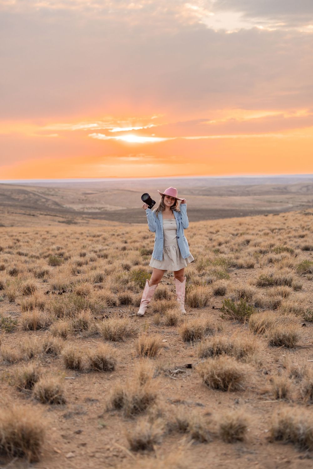 Woman photographer in denim jacket and pink hat holds camera in desert landscape at golden sunset.