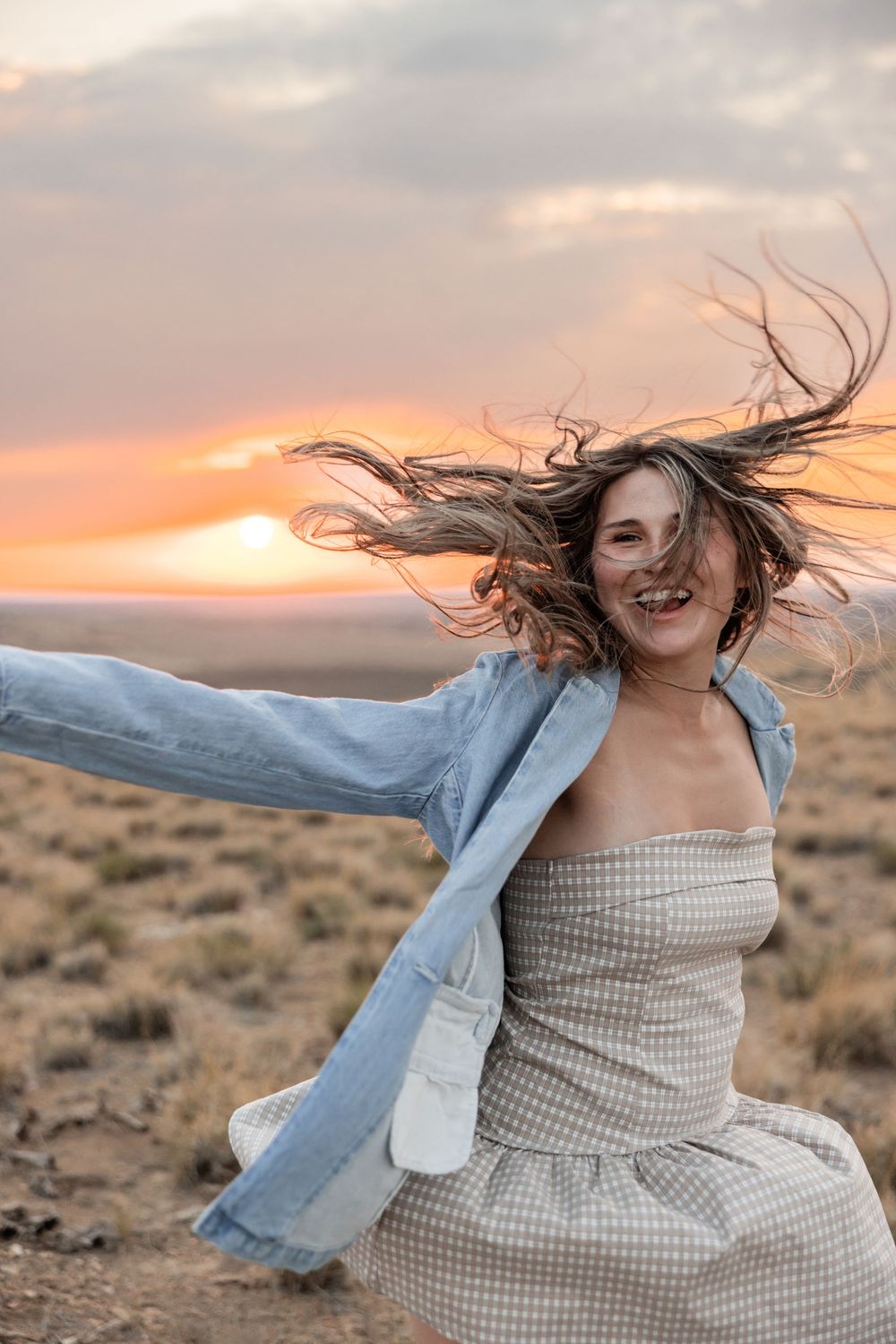Joyful woman in gingham dress and denim jacket with wind-blown hair dancing at sunset in open desert landscape.