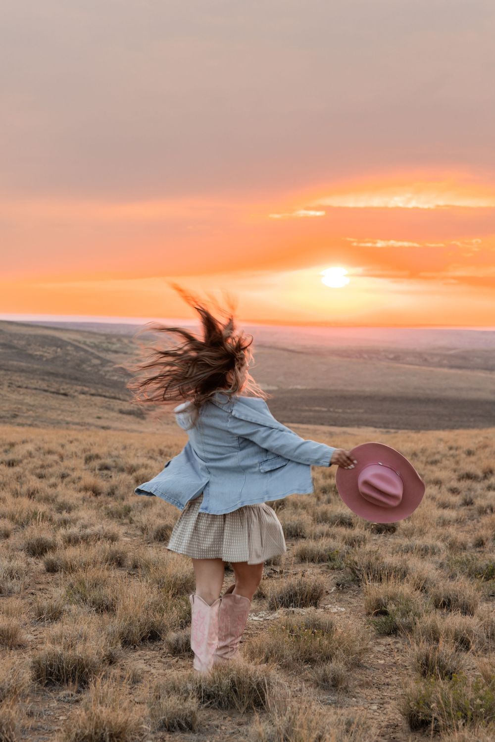 A woman twirls in a field at sunset, wearing a blue jacket, skirt, pink boots, holding a pink cowboy hat.