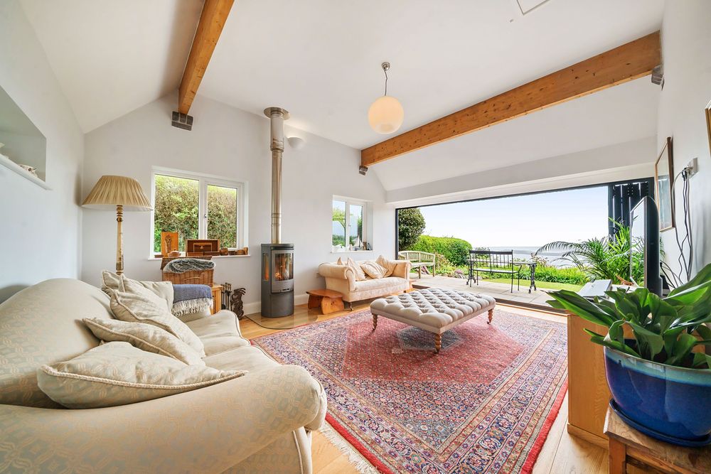Bright living room with exposed wooden beams, wood burner, large sliding doors, red Persian rug, and scenic garden views.