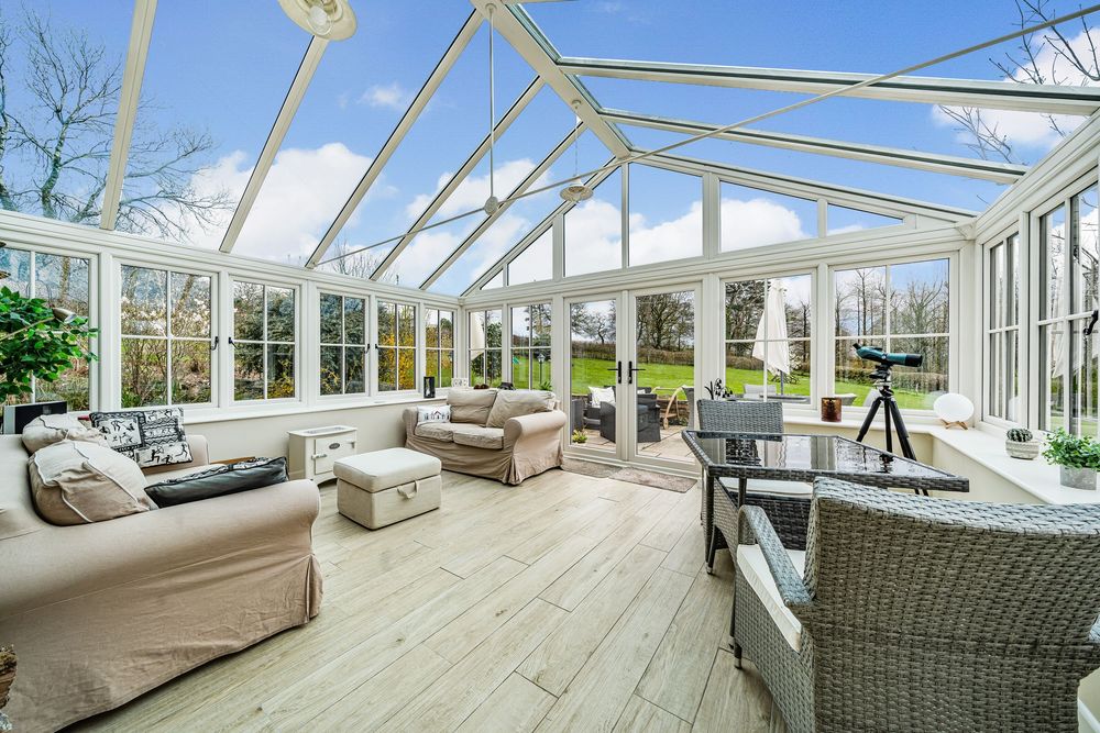 Bright conservatory with vaulted glass roof, cream sofas, wicker chairs, and hardwood floors overlooking a lush green garden.