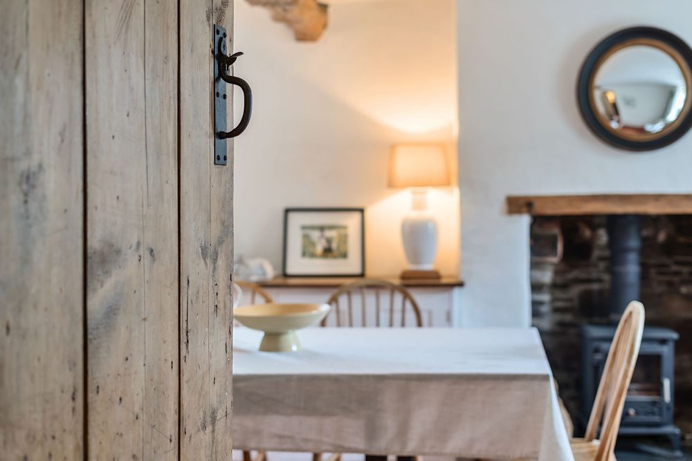 Rustic wooden door opens to a cozy cottage dining room with stone fireplace, linen tablecloth, and warm lamp light.