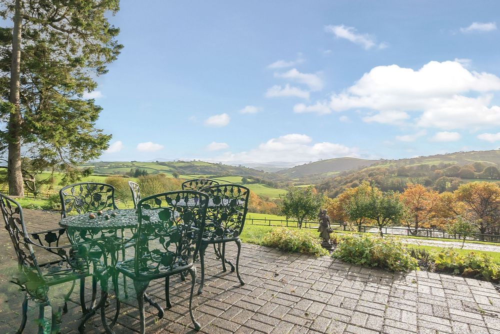 Green wrought iron patio furniture on stone terrace overlooking lush rolling hills and valleys under a bright blue sky.