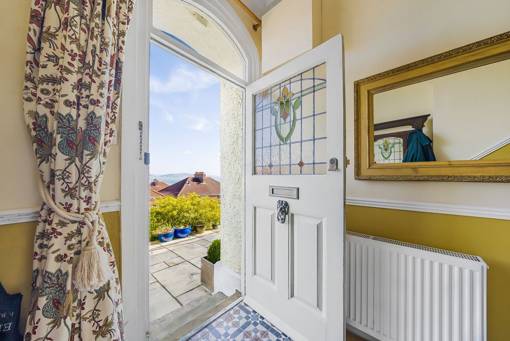 A bright hallway with an open white front door featuring stained glass, yellow walls, floral curtains, and a gold-framed mirror.