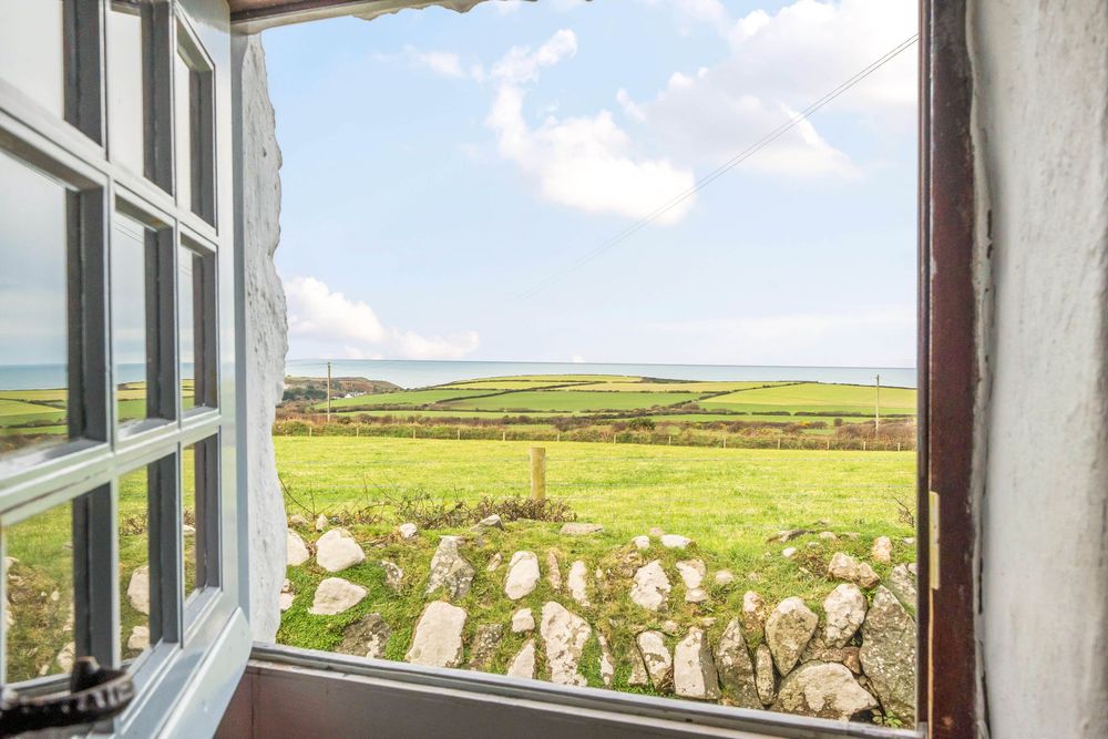 Open window with a stunning rural view of green fields, stone walls, and a glimpse of the sea under a blue sky.