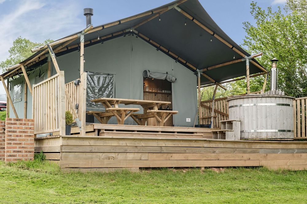 Safari tent with wooden deck, hot tub, picnic table, and log burner chimney surrounded by lush green gardens.