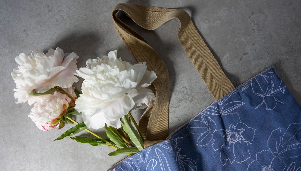 2 white peonies lying inside a blue totebag with a white hand drawn flower botanical