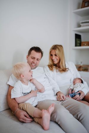 Family relaxes on white couch with toddler and newborn in casual home setting.