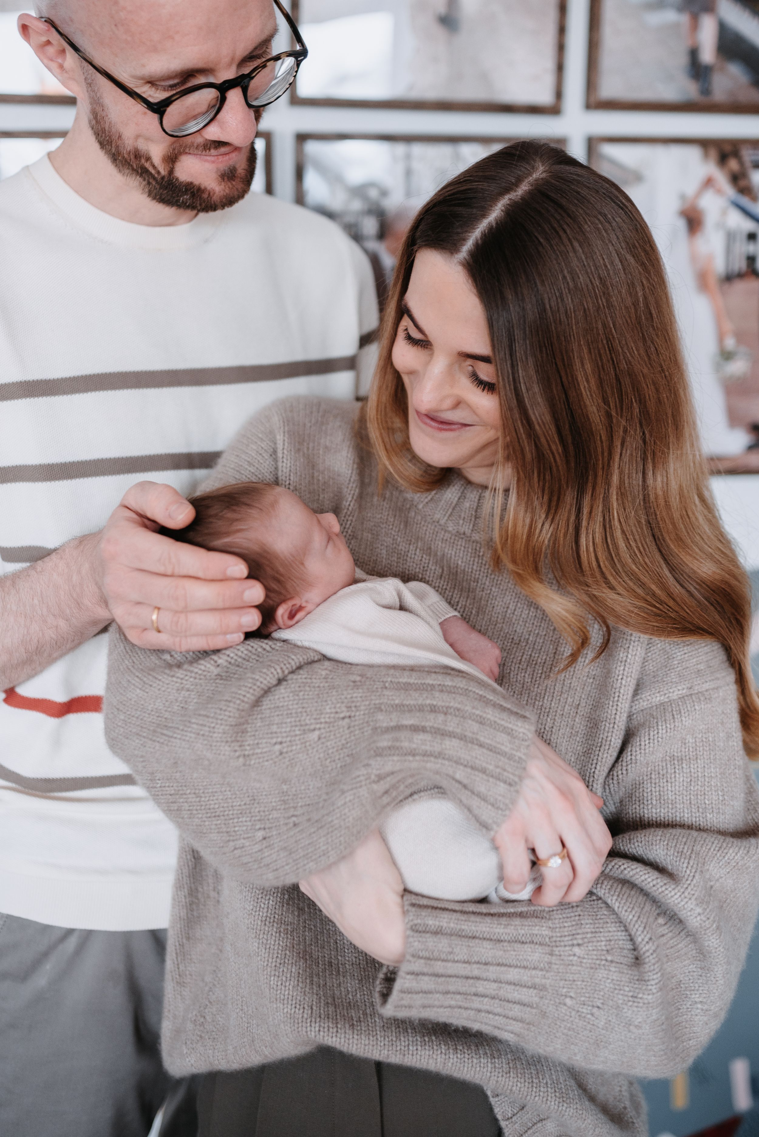 Couple in cozy sweaters holds and gazes lovingly at their sleeping newborn baby in their arms.