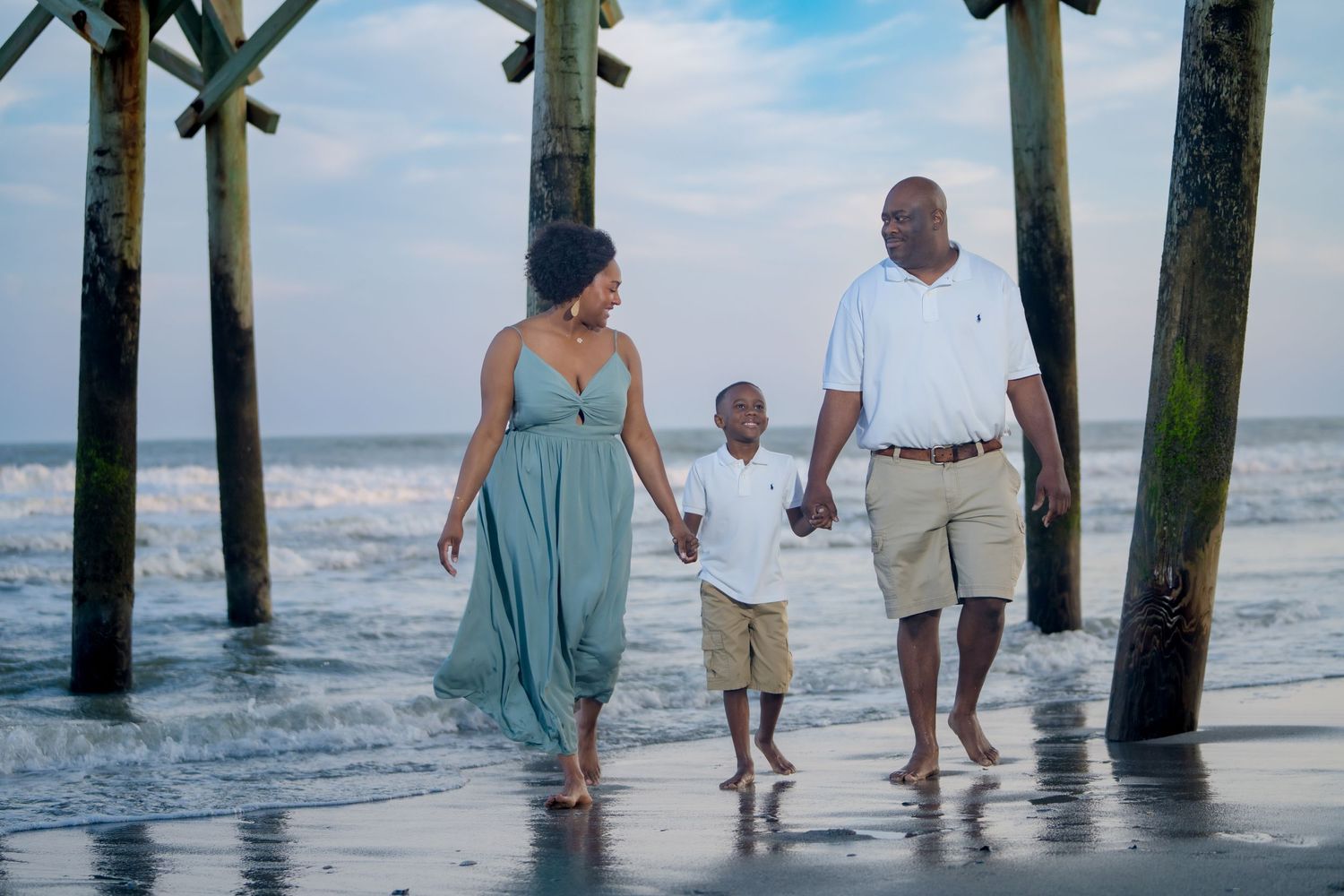 A family walks together between pier pillars on a beach at sunset.