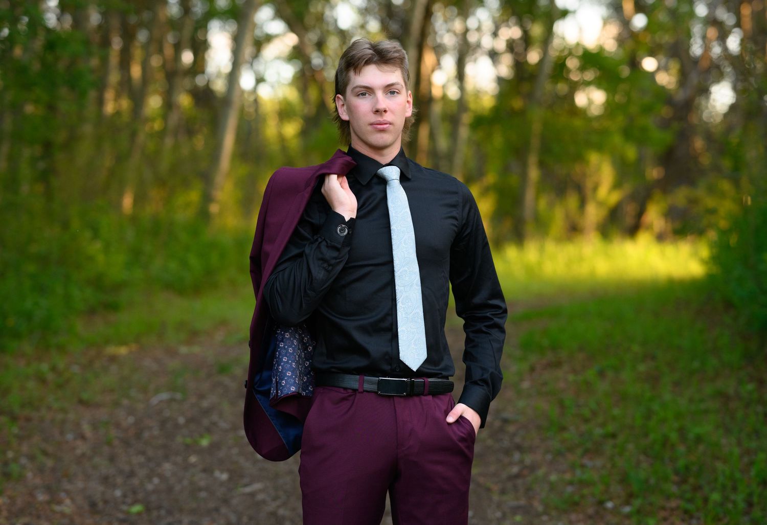 A formal portrait of someone in a burgundy suit and black shirt standing on a wooded path at sunset.